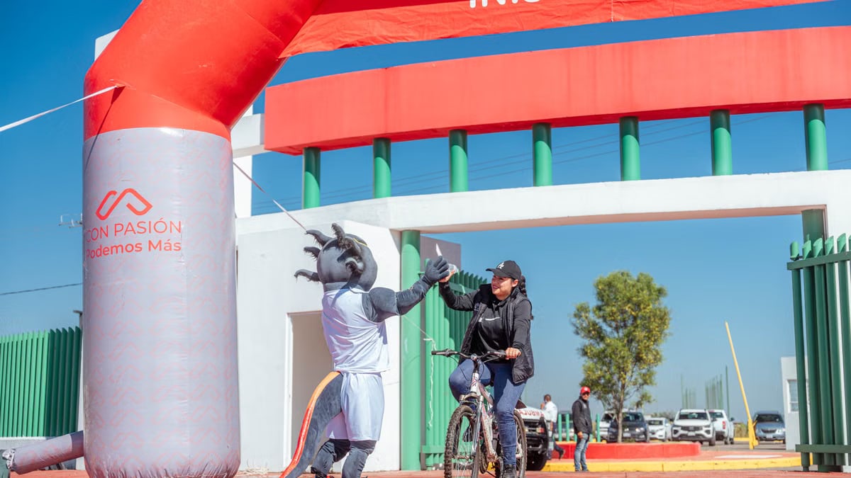A cyclist receives a high-five from a mascot at the finish line of a cycling event under a clear blue sky.